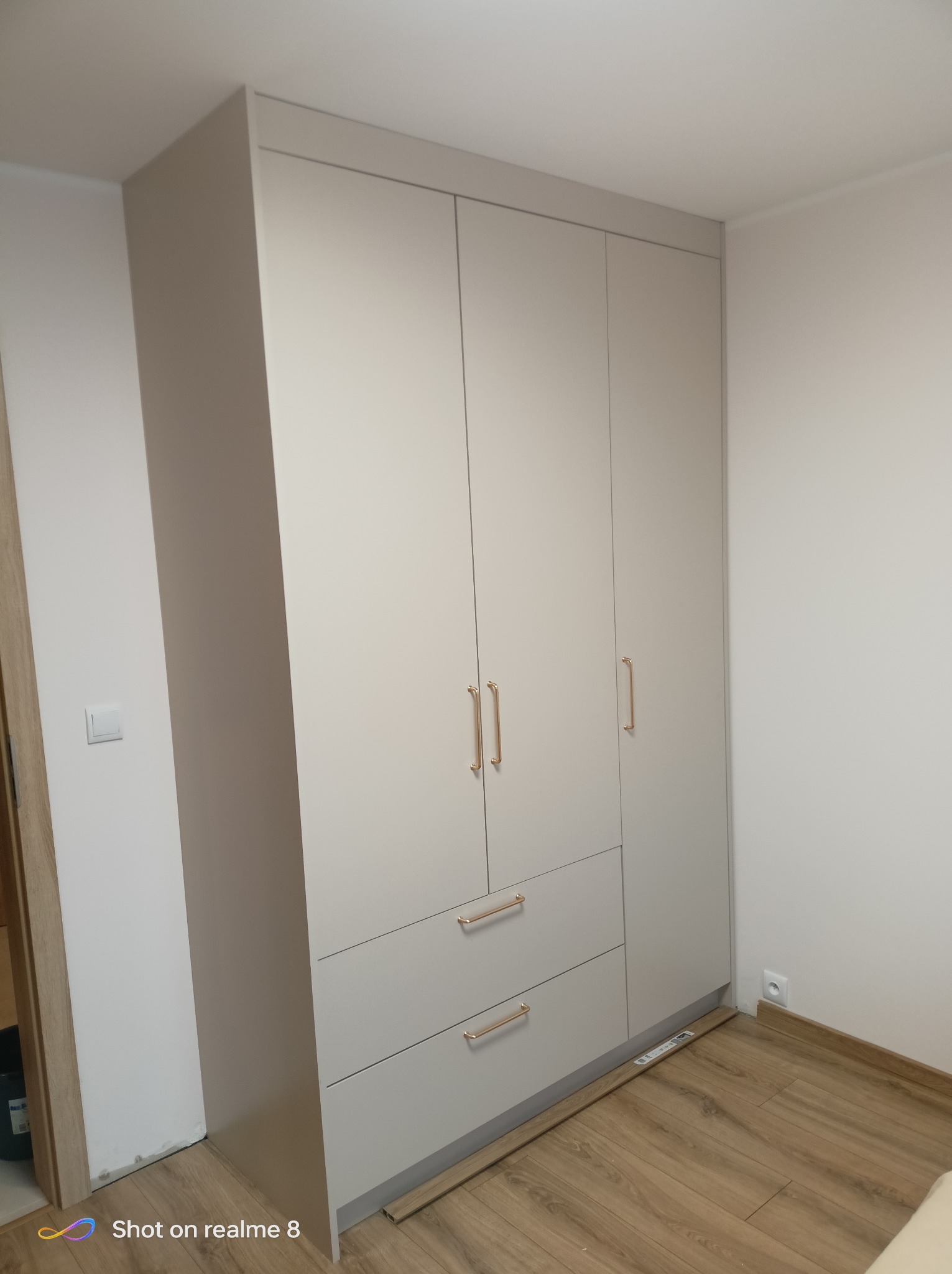 Jasnoszara szafa trzydrzwiowa z szufladami i złotymi uchwytami, dopasowana do wnętrza pokoju z drewnianą podłogą. Widoczny fragment ściany z gniazdkiem elektrycznym.