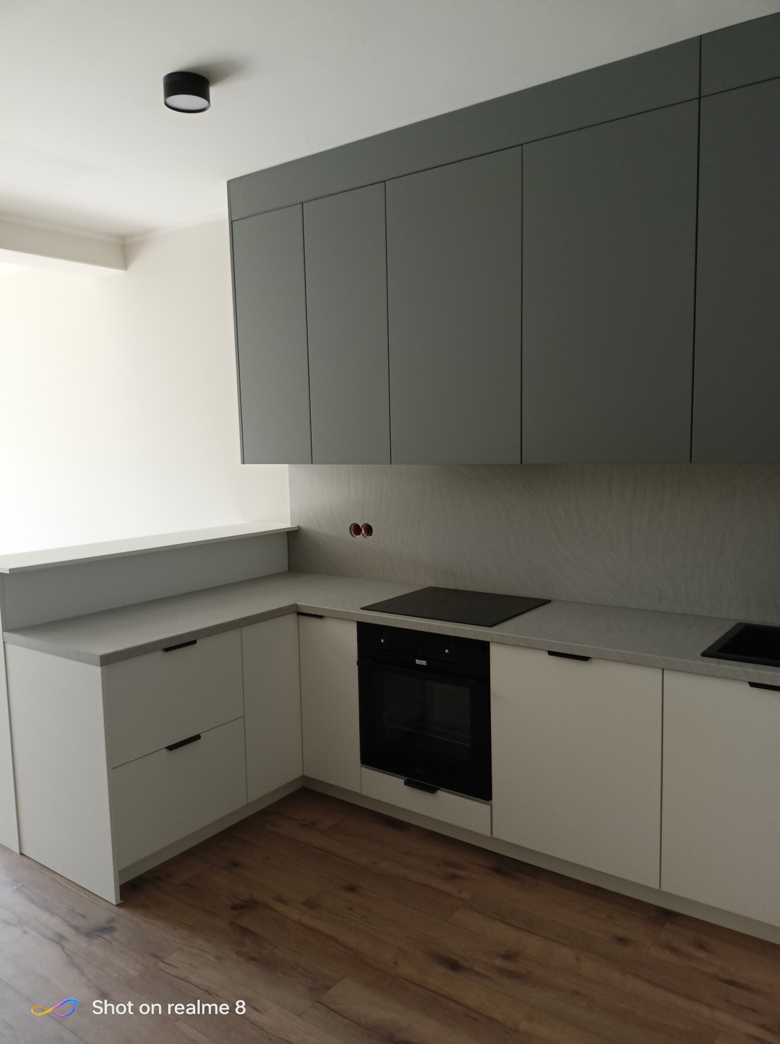 Nowoczesna kuchnia z szarymi szafkami górnymi i białymi dolnymi, drewniana podłoga. Minimalistyczny design, czarne uchwyty, płyta indukcyjna. Oświetlenie sufitowe.