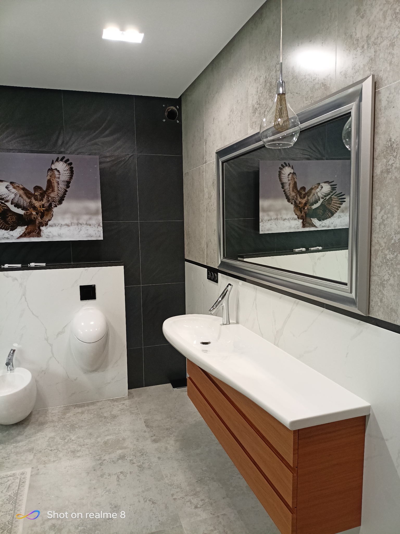 Nowoczesna łazienka z umywalką na drewnianej szafce, bidetem i pisuarami. Na ścianie obraz z orłem, odbity w lustrze. Szare płytki podłogowe i oświetlenie punktowe.