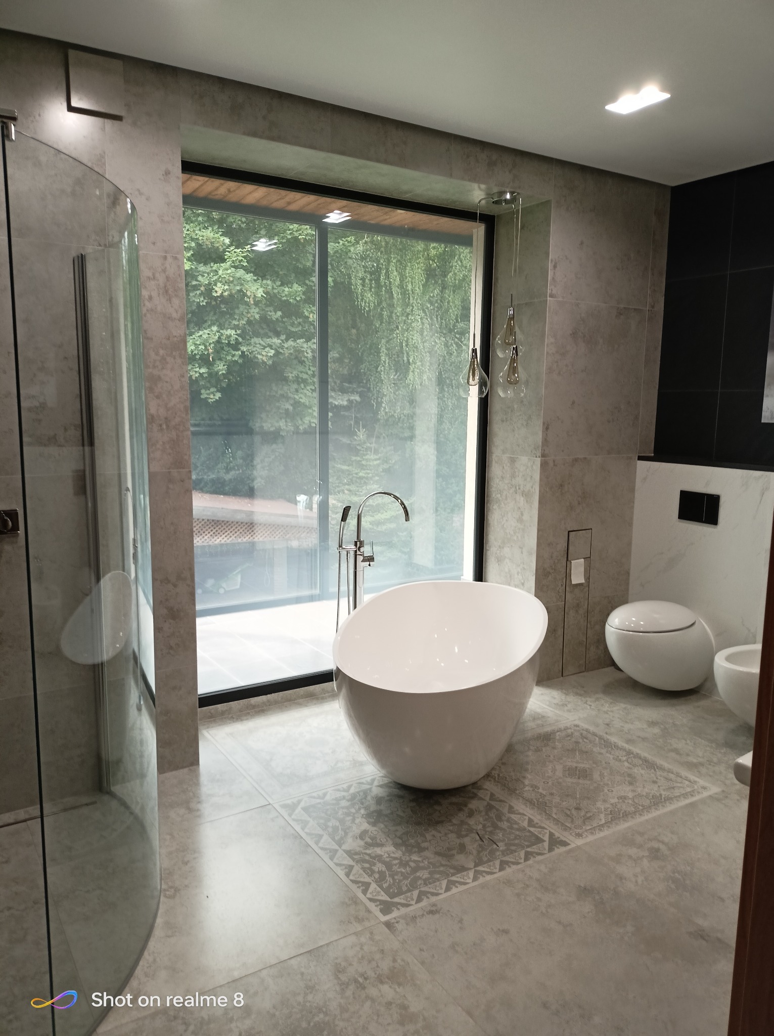 Nowoczesna łazienka z wolnostojącą wanną na tle okna z widokiem na zieleń. Szare płytki imitujące kamień, minimalistyczna armatura i oświetlenie.