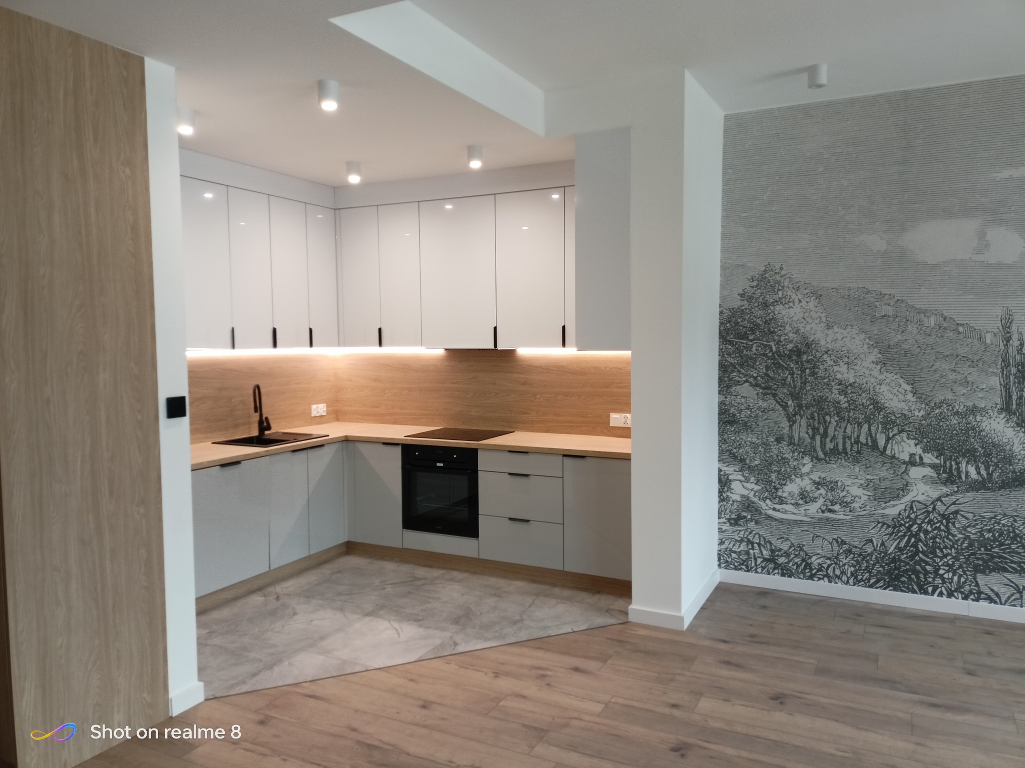 Nowoczesna kuchnia w odcieniach bieli i drewna, z subtelnym oświetleniem LED pod szafkami i dekoracyjną tapetą z motywem pejzażu na ścianie.
