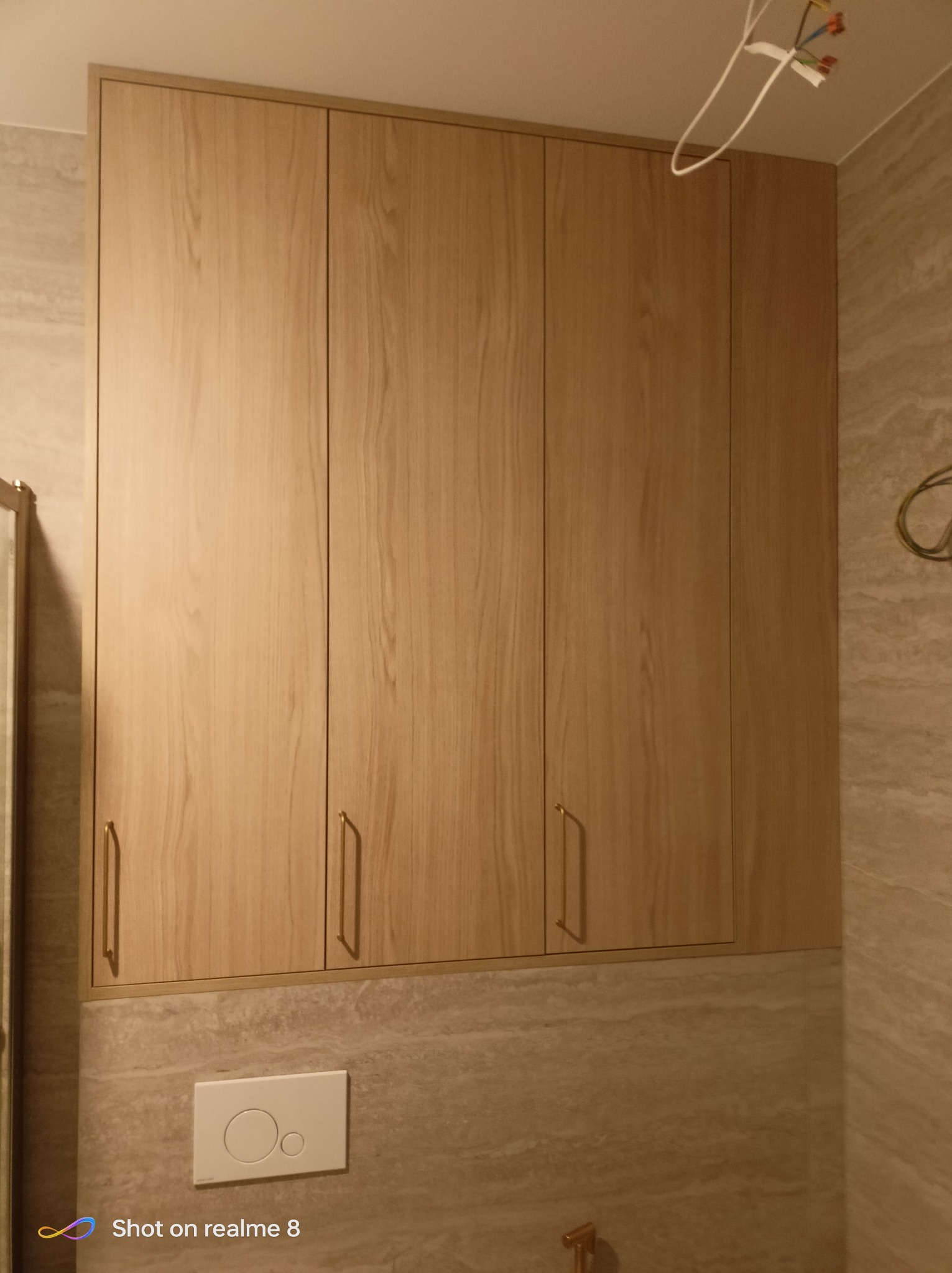 Drewniana szafka łazienkowa z czterema drzwiczkami i złotymi uchwytami, zamontowana nad spłuczką toalety, na tle marmurowej ściany. Widoczne przewody elektryczne.