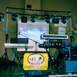 Scena muzyczna z dwoma keyboardami, nagłośnieniem, oświetleniem i wyświetlanym logo zespołu 'Magnes' na ekranie w tle.