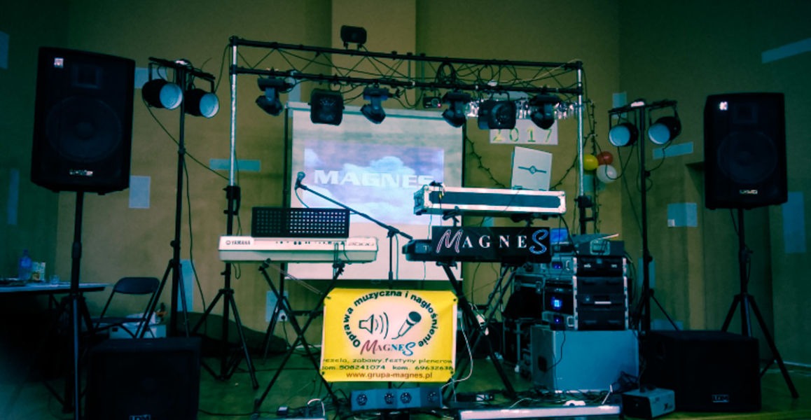 Scena muzyczna z dwoma keyboardami, nagłośnieniem, oświetleniem i wyświetlanym logo zespołu 'Magnes' na ekranie w tle.