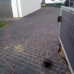 Ułożona, ciemnoszara kostka na podjeździe z widoczną fugą piaskową, obok domu z białą elewacją i metalową bramą.