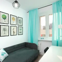 Jasne wnętrze pokoju z szarą sofą, białym biurkiem i turkusowymi zasłonami, ozdobione sześcioma grafikami w czarnych ramkach na ścianie.