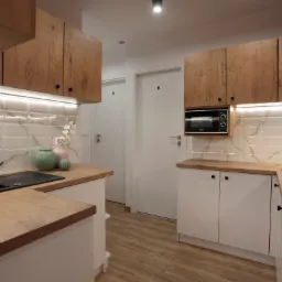 Nowoczesna kuchnia z białymi szafkami dolnymi i drewnopodobnymi górnymi, podświetlane płytki imitujące marmur, czarny zlew i bateria, drewniana podłoga.