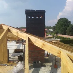 Wznoszenie komina z czarnej cegły klinkierowej na dachu w trakcie budowy, widoczne drewniane elementy konstrukcji dachu i fragmenty muru z czerwonej cegły.