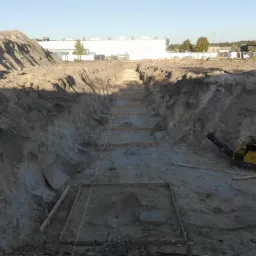 Wykop pod fundamenty z drewnianym szalunkiem i ubijarką wibracyjną na dnie, widok z góry na plac budowy z pryzmą ziemi w tle.