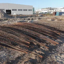 Siatka zbrojeniowa ułożona na piaszczystym placu budowy, w tle widoczny budynek przemysłowy i hałda piasku.