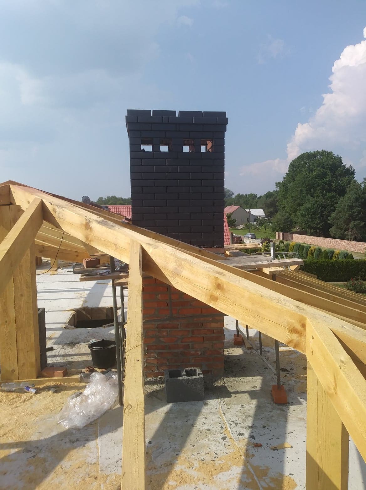 Wznoszenie komina z czarnej cegły klinkierowej na dachu w trakcie budowy, widoczne drewniane elementy konstrukcji dachu i fragmenty muru z czerwonej cegły.