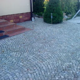 Ułożona z szarej kostki granitowej nawierzchnia w kształcie wachlarza, prowadząca do schodów wejściowych budynku, z widocznym pracownikiem w tle.