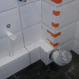 Białe płytki ścienne z pomarańczowymi zabezpieczeniami narożników, czarne płytki podłogowe, biała rura odpływowa i szara rura kanalizacyjna w trakcie montażu w łazience.