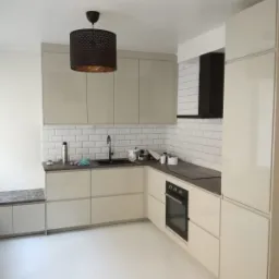 Jasna kuchnia z białymi, prostokątnymi płytkami na ścianie, czarnym okapem i czarną armaturą, oświetlona lampą z ażurowym abażurem