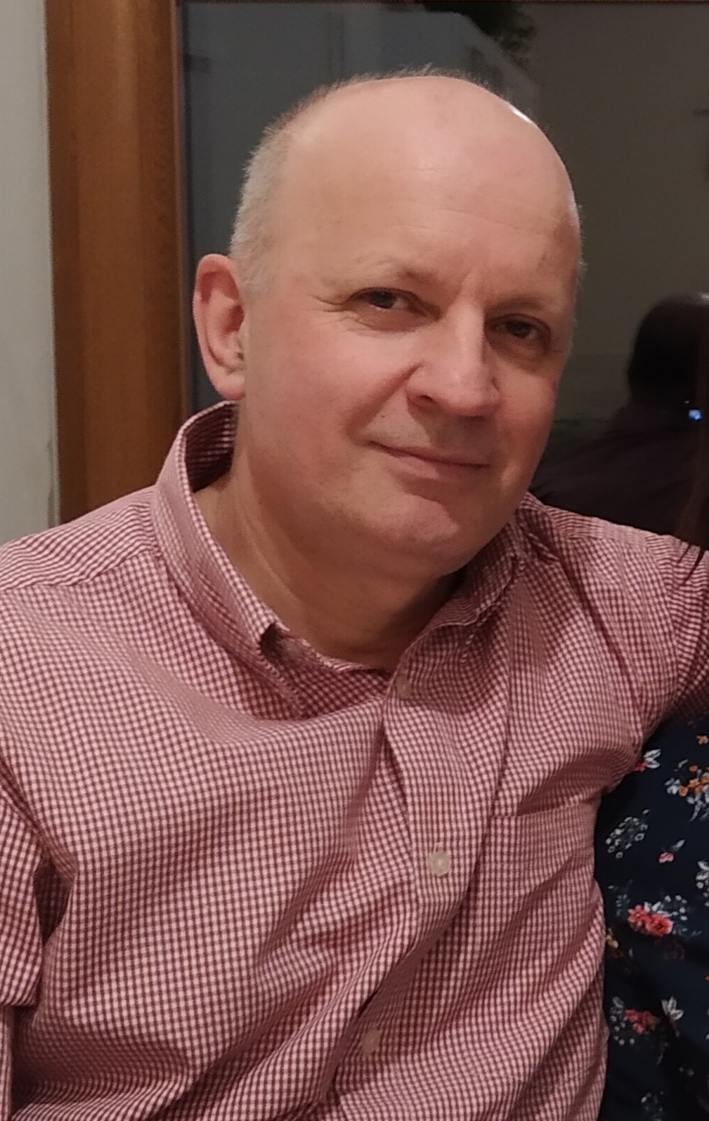 Portret mężczyzny w koszuli w drobną kratę, z łagodnym uśmiechem, na neutralnym tle. Widoczny fragment ramienia drugiej osoby.