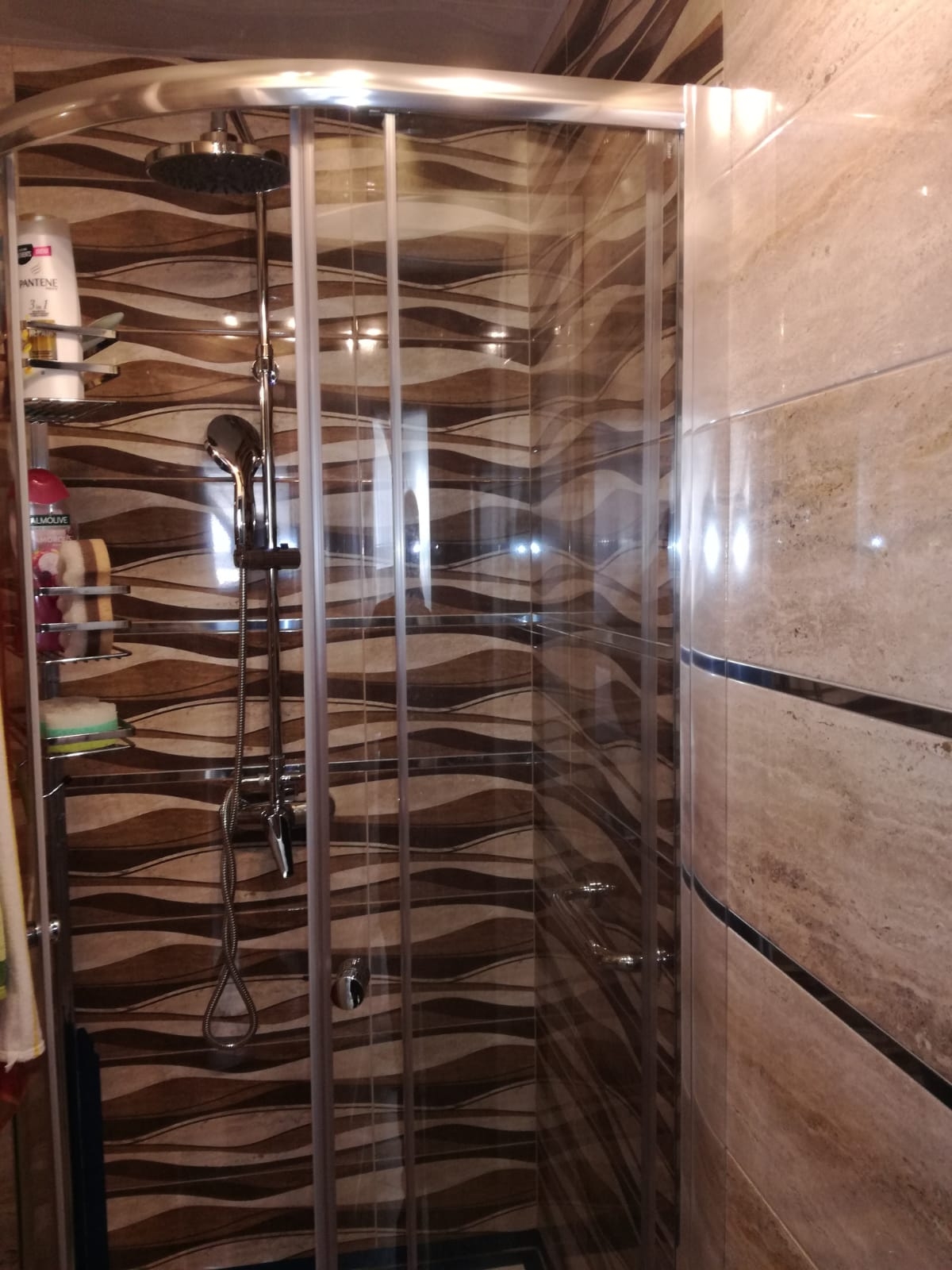 Kabina prysznicowa z chromowanymi elementami i półkami w łazience wyłożonej płytkami w odcieniach brązu i beżu z motywem fal