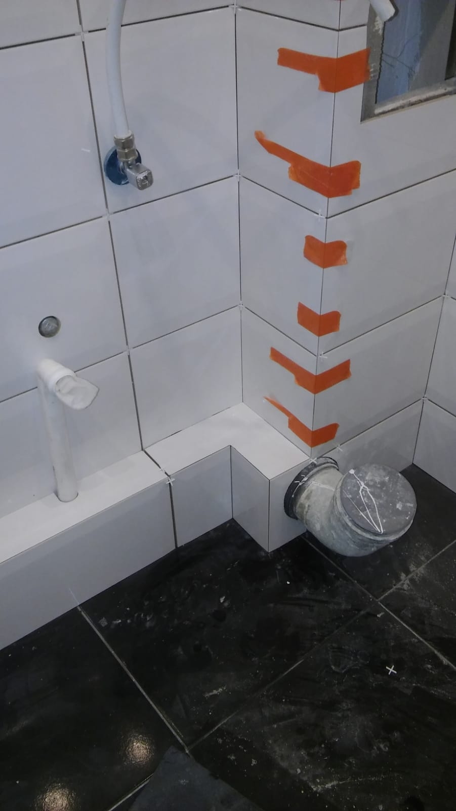 Białe płytki ścienne z pomarańczowymi zabezpieczeniami narożników, czarne płytki podłogowe, biała rura odpływowa i szara rura kanalizacyjna w trakcie montażu w łazience.