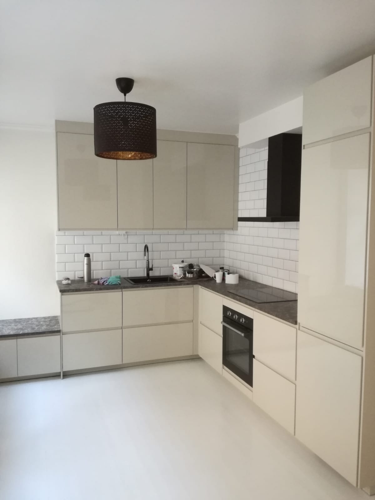 Jasna kuchnia z białymi, prostokątnymi płytkami na ścianie, czarnym okapem i czarną armaturą, oświetlona lampą z ażurowym abażurem