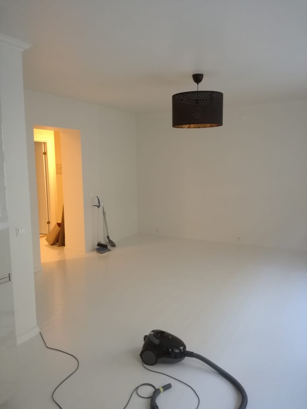 Pomalowane na biało puste pomieszczenie po remoncie, z odkurzaczem na podłodze, widoczne narzędzia malarskie i czarna lampa wisząca.