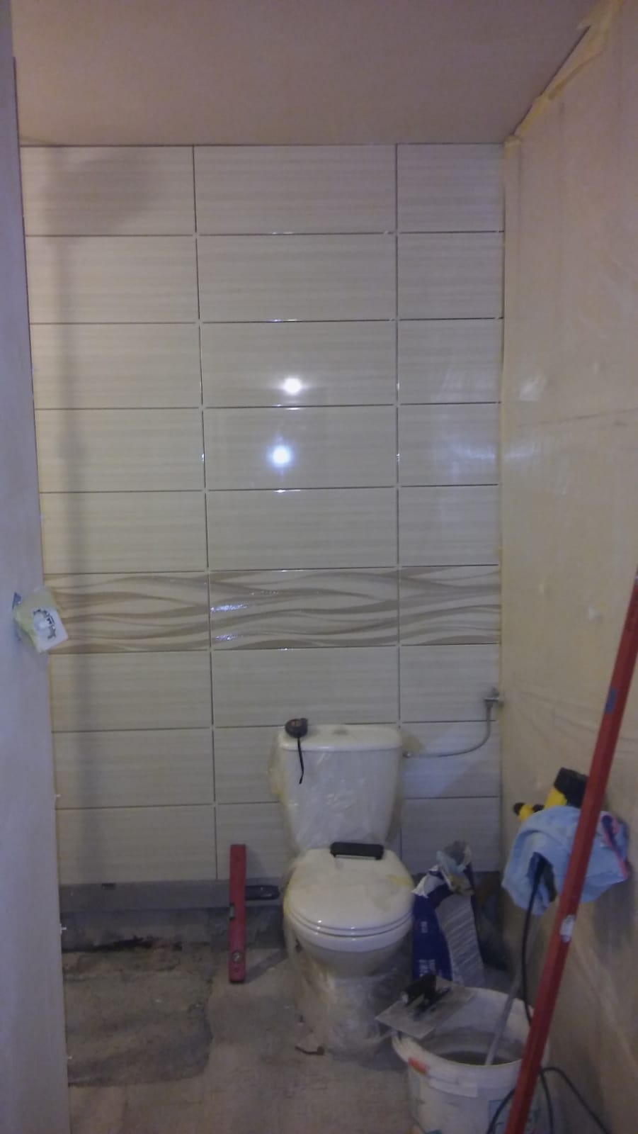 Ściana w łazience z ułożonymi beżowymi płytkami z poziomym wzorem fal, częściowo zakryty sedes w folii ochronnej, poziomica i wiadro z zaprawą na podłodze w trakcie prac wykończeniowych.