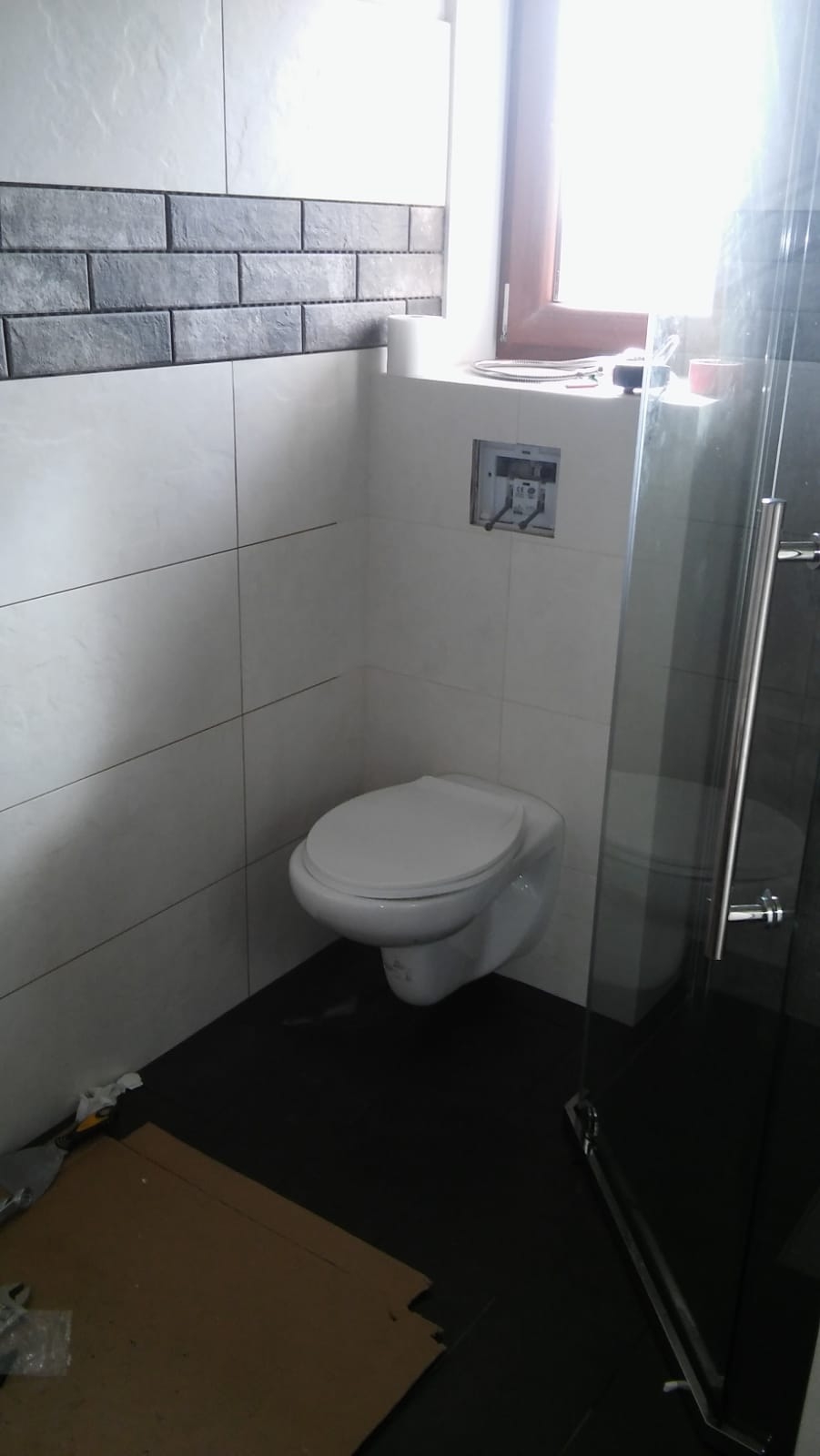 Świeżo wykończona łazienka z wiszącą toaletą, ciemną podłogą i szarymi płytkami imitującymi cegłę na ścianie, widoczna instalacja podtynkowa.