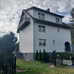 Dwupiętrowy dom z balkonem, świeżo po malowaniu elewacji na biało, widoczny fragment ogrodzenia i osoba przygotowująca się do malowania w tle, Bolesławiec.