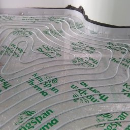 PANEA - Białe rury ogrzewania podłogowego ułożone w pętle na srebrzystej folii izolacyjnej Kingspan Therma, widok z góry, etap instalacji.