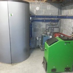 PANEA - Szara akumulacyjna jednostka grzewcza i zielony piec na paliwo stałe w kotłowni z instalacjami na ścianie z betonowych bloków.