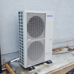 PANEA - Biała zewnętrzna jednostka klimatyzacji Samsung Digital Inverter zamontowana na betonowej podstawie przy szarej ścianie budynku, widoczne rury i elementy instalacji, w tle gruz i siatka zbrojeniowa.