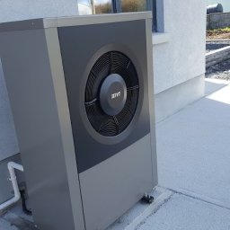 PANEA - Szara pompa ciepła IVT zamontowana na zewnątrz budynku, widoczne białe rury instalacyjne oraz częściowo zagospodarowany teren w tle.