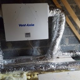 PANEA - Centrala wentylacyjna Vent-Axia z aluminiowymi przewodami w szarej przestrzeni poddasza w Kamionnej, widoczne elementy konstrukcji drewnianej dachu.