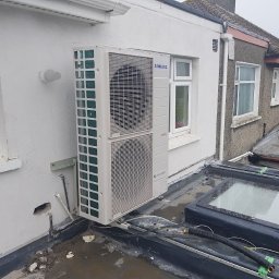 PANEA - Biała, dwuczęściowa jednostka zewnętrzna klimatyzacji marki Samsung zamontowana na wspornikach na płaskim dachu budynku z oknami dachowymi, widoczne przewody i instalacja elektryczna.