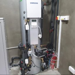 PANEA - Szara, pionowa pompa ciepła Samsung z czarnymi rurami i przewodami w surowym, betonowym pomieszczeniu technicznym.