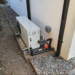 PANEA - Biała pompa ciepła zainstalowana na betonowej podstawie przy ścianie budynku, z widocznymi rurami i zaworami w kolorze czarnym i czerwonym. W tle żwir i fragment rynny.