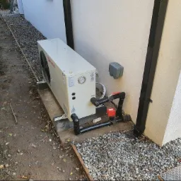 Biała pompa ciepła zainstalowana na betonowej podstawie przy ścianie budynku, z widocznymi rurami i zaworami w kolorze czarnym i czerwonym. W tle żwir i fragment rynny.