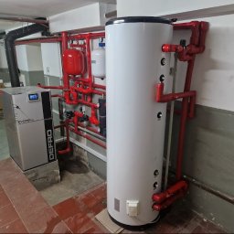 PANEA - Kotłownia z kotłem na pellet Defro Evopell i białym zasobnikiem ciepłej wody, rury w kolorze czerwonym, płytki podłogowe w odcieniach brązu, widoczne elementy instalacji grzewczej.