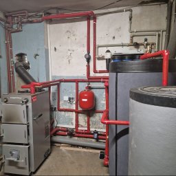 PANEA - Szary piec Defro z podłączeniem do instalacji grzewczej z czerwonymi rurami i dwoma szarymi zbiornikami w piwnicy z betonowymi ścianami.