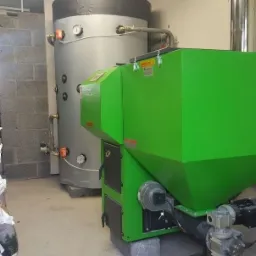 Zielony piec na pellet z zasobnikiem obok srebrnego zbiornika buforowego, obok sterta worków z pelletem w pomieszczeniu kotłowni.