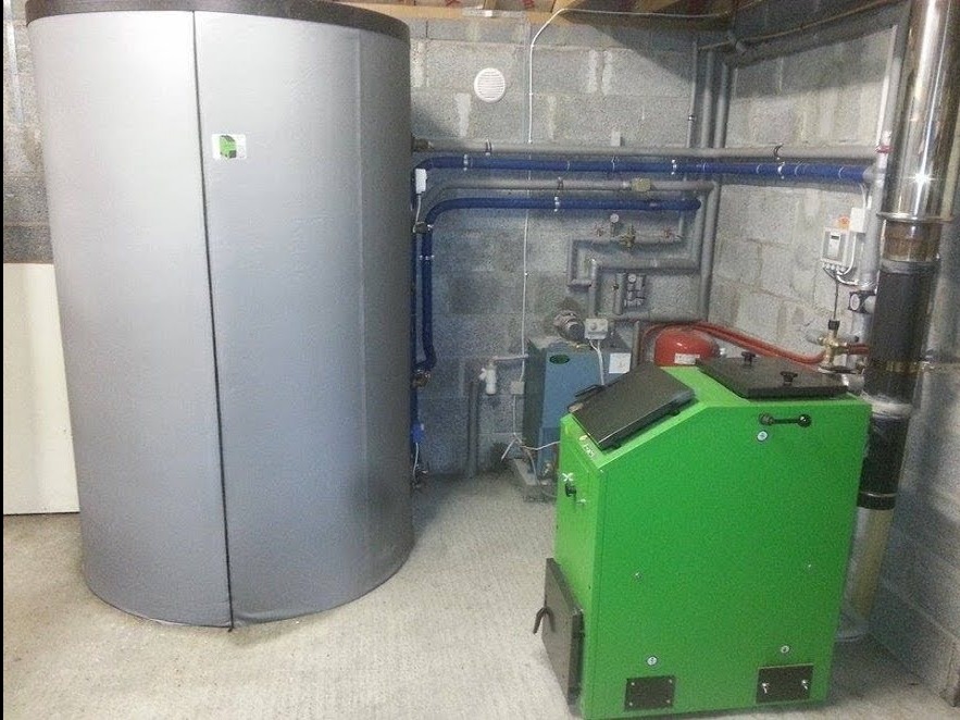 Szara akumulacyjna jednostka grzewcza i zielony piec na paliwo stałe w kotłowni z instalacjami na ścianie z betonowych bloków.