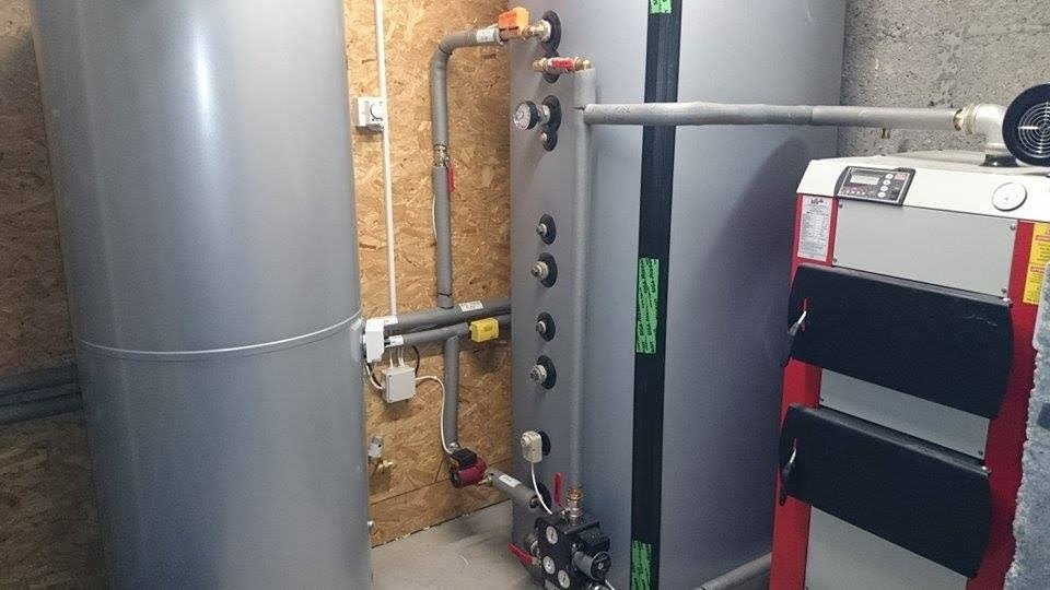 Szare zbiorniki akumulacyjne ciepła z rurami i osprzętem w kotłowni, widoczny kocioł grzewczy z czarnymi drzwiczkami.