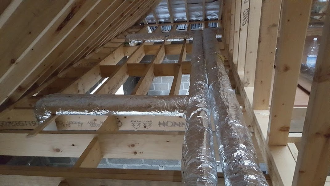 Instalacja przewodów wentylacyjnych w przestrzeni poddasza, widoczne drewniane krokwie i folia izolacyjna na rurach.