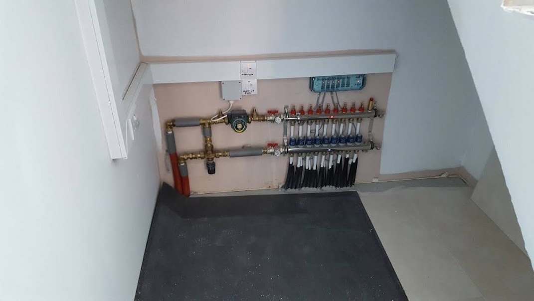 Rozdzielacz ogrzewania podłogowego z licznymi pętlami, zamontowany w rogu pomieszczenia ze skosami, widoczny na tle beżowej ściany z szarą matą pod spodem.