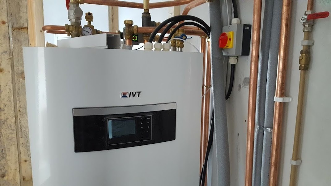 Biała pompa ciepła IVT z widocznym panelem sterowania, miedzianymi rurami i wyłącznikiem awaryjnym na ścianie.