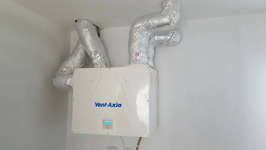 Centrala wentylacyjna Vent-Axia zamontowana na ścianie z podłączonymi przewodami wentylacyjnymi w izolacji aluminiowej.