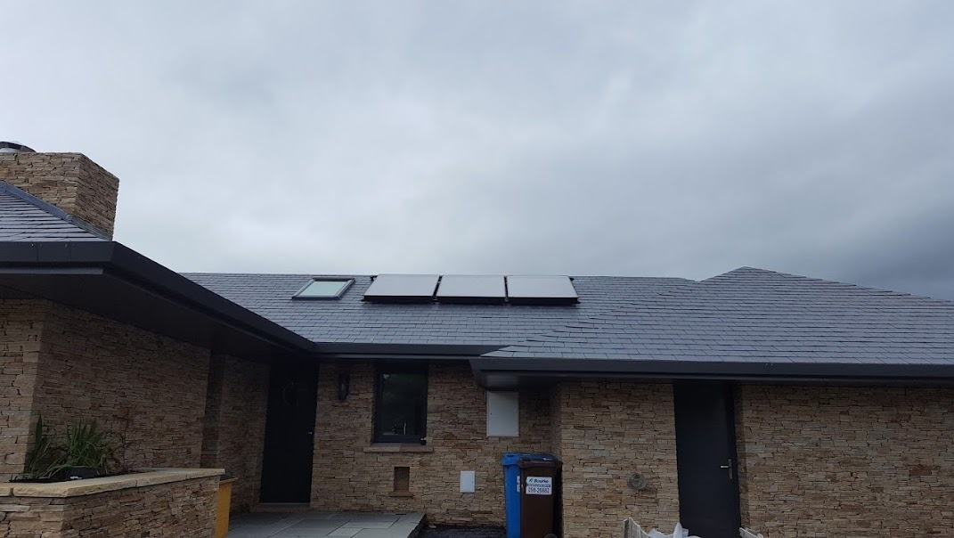 Dom jednorodzinny z panelami fotowoltaicznymi na dachu pokrytym szarym łupkiem, widoczne okno dachowe i komin obłożony kamieniem, pochmurne niebo w tle.