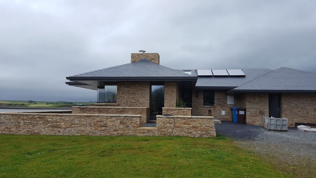 Dom jednorodzinny z kamienną elewacją, szarym dachem z panelami fotowoltaicznymi i widokiem na krajobraz za ogrodzeniem.