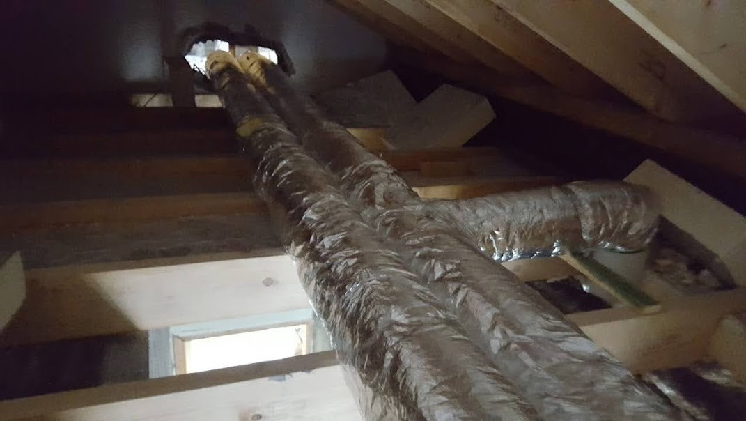 Dwie rury wentylacyjne w izolacji aluminiowej, prowadzone przez drewnianą konstrukcję poddasza, widoczne elementy ocieplenia