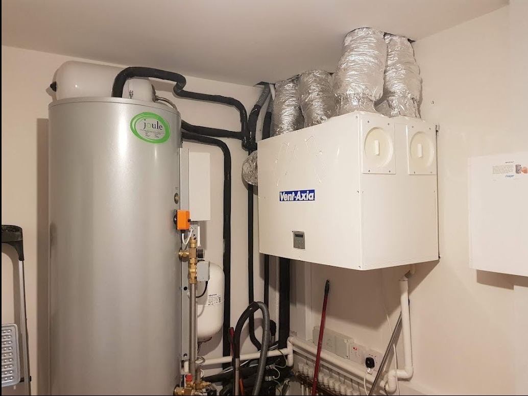 Srebrny zasobnik ciepłej wody użytkowej Joule z zielonym logo, podłączony do instalacji, obok biała centrala wentylacyjna Vent-Axia z aluminiowymi przewodami wentylacyjnymi, widoczne rury...