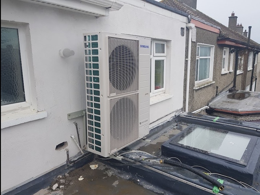 Biała, dwuczęściowa jednostka zewnętrzna klimatyzacji marki Samsung zamontowana na wspornikach na płaskim dachu budynku z oknami dachowymi, widoczne przewody i instalacja elektryczna.