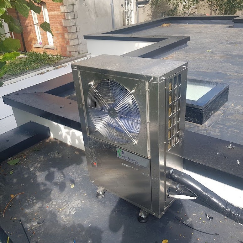 Błyszcząca, metalowa pompa ciepła z wentylatorem, umieszczona na płaskim dachu z widocznym świetlikiem dachowym i fragmentem budynku w tle.